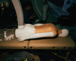 Mannequin auf Liege, Handschuhe, Kabel, Person im Hintergrund.