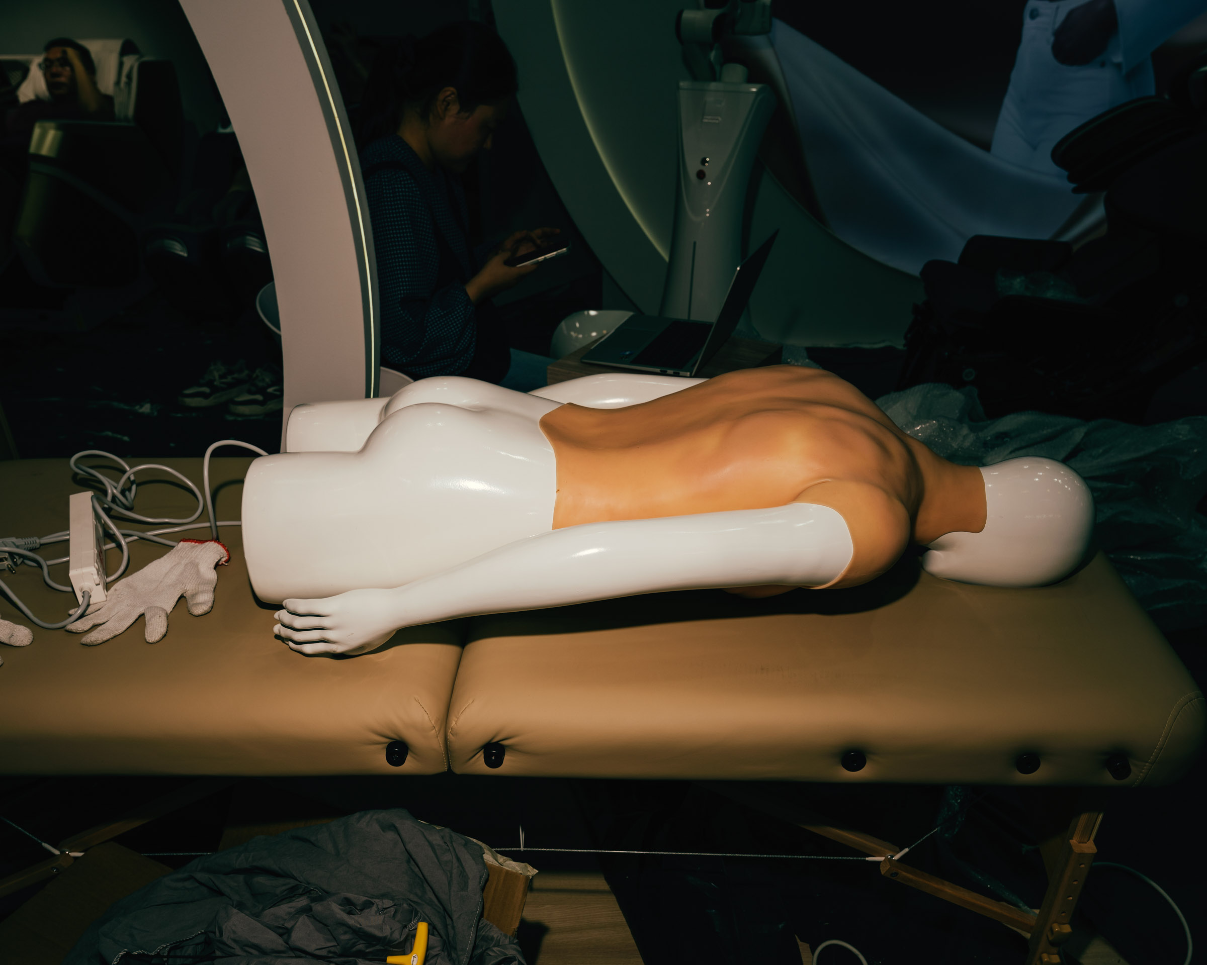 Mannequin auf Liege, Handschuhe, Kabel, Person im Hintergrund. 