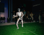 Roboter auf Kunstrasen mit Zuschauern im Hintergrund.