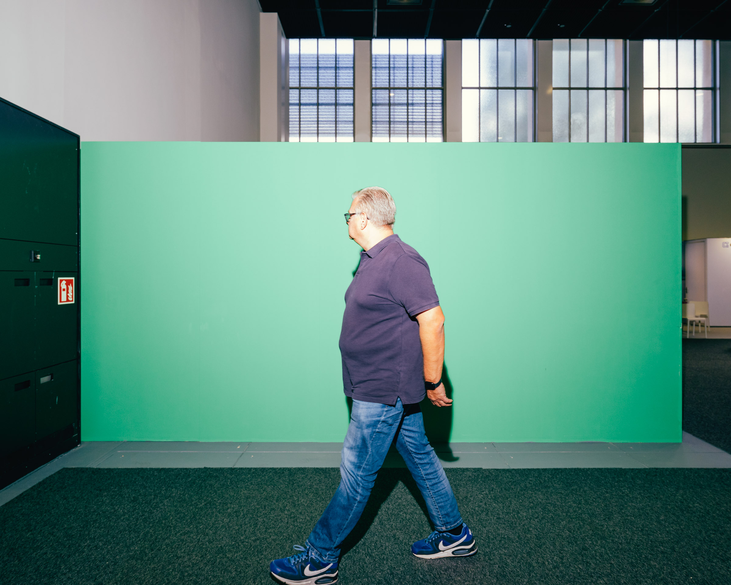 Mann geht vor grüner Wand, blaue Jeans, blaue Schuhe. 
